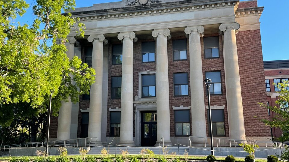 Image of Louise Pound Hall, 3 story red brick and white sandstone building, Classical Revival style, Southern exposure, sunny day, leafed-out tree on the left, bright blue sky above